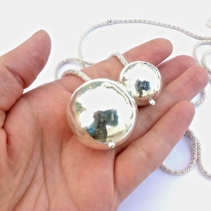 May include: A silver necklace with two round, smooth, silver pendants. The necklace is made of a fine chain.