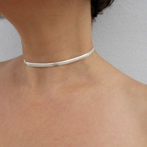 May include: A silver-toned snake chain choker necklace featuring a flat, flexible, and tightly woven link design that sits closely around the neck. The metallic surface has a polished finish that reflects light, creating a smooth and continuous appearance.