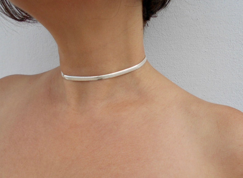May include: A silver-toned snake chain choker necklace featuring a flat, flexible, and tightly woven link design that sits closely around the neck. The metallic surface has a polished finish that reflects light, creating a smooth and continuous appearance.