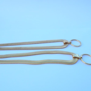 May include: Two gold-colored necklaces with a herringbone chain design. Each necklace features a rectangular clasp and a circular pendant. The necklaces are displayed against a light blue background.