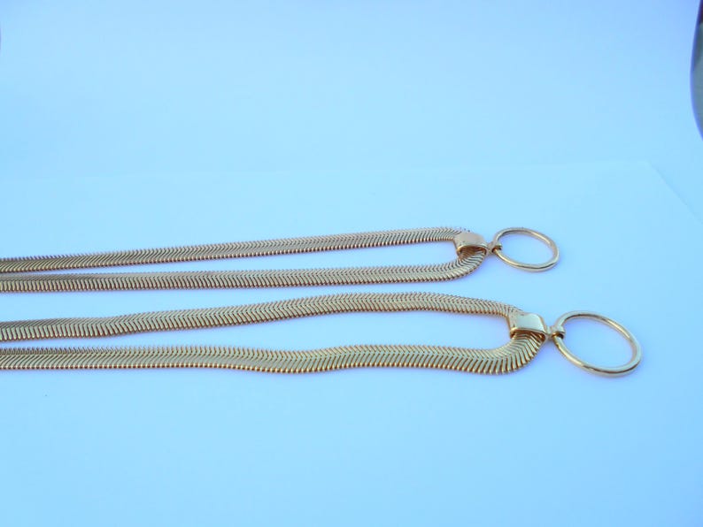 May include: Two gold-colored necklaces with a herringbone chain design. Each necklace features a rectangular clasp and a circular pendant. The necklaces are displayed against a light blue background.
