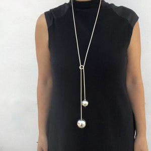 May include: A long silver chain necklace with two large silver spheres hanging from it. The necklace is worn by a person wearing a black sleeveless top.