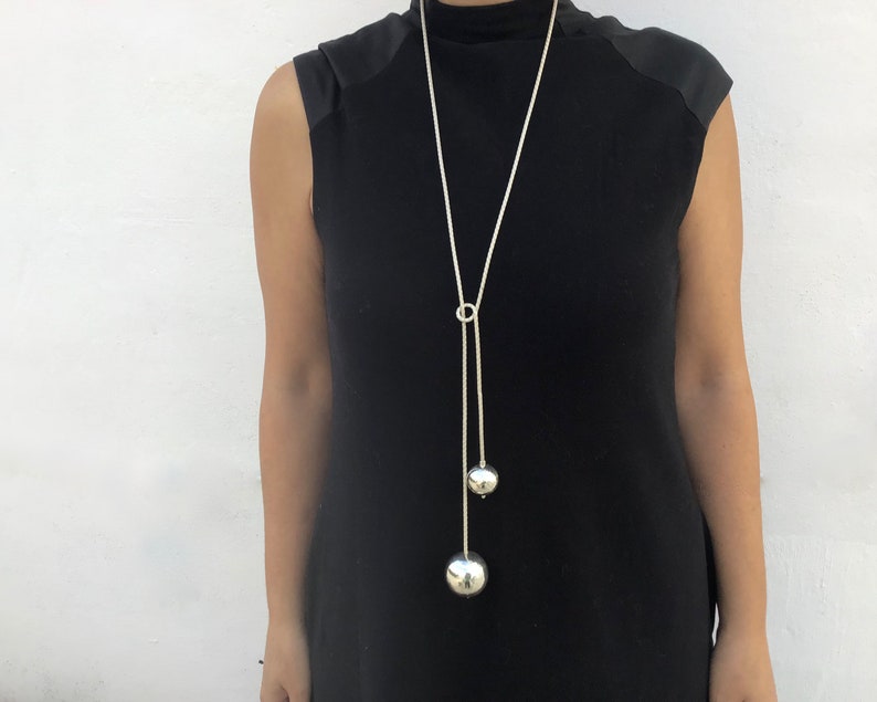 May include: A long silver chain necklace with two large silver spheres hanging from it. The necklace is worn by a person wearing a black sleeveless top.