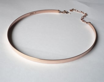 May include: A rose gold choker necklace with a simple, flat design. The necklace has a clasp closure and is made of metal.
