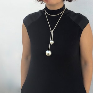 May include: A silver necklace with two large, round, silver charms. The necklace is on a model wearing a black mock neck shirt.