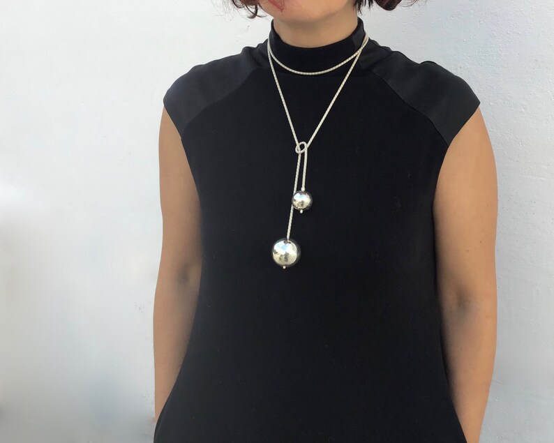 May include: A silver necklace with two large, round, silver charms. The necklace is on a model wearing a black mock neck shirt.