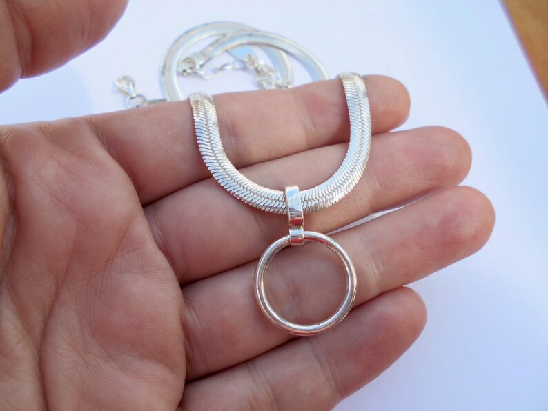 May include: A silver-toned herringbone chain necklace featuring a smooth, circular pendant attached to a polished bail. The flat, woven links of the chain create a sleek, reflective surface that leads to the simple, open ring charm at the center.