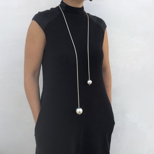 May include: A long silver chain necklace with two large silver spheres hanging from it. The necklace is worn by a person wearing a black mock neck shirt.