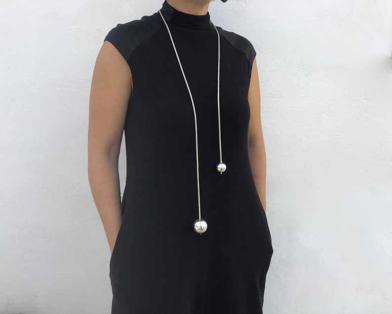 May include: A long silver chain necklace with two large silver spheres hanging from it. The necklace is worn by a person wearing a black mock neck shirt.