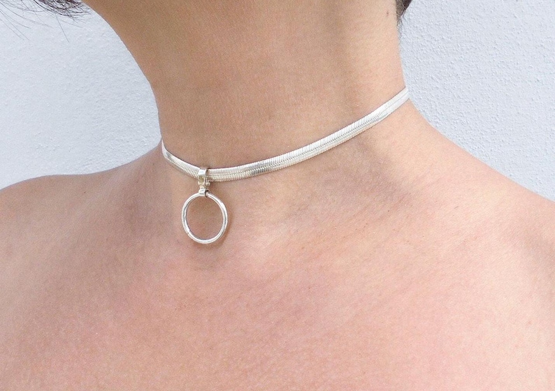May include: A silver-toned choker necklace featuring a flat, flexible herringbone chain design. The necklace includes a central circular ring pendant attached to the chain by a small, fixed metal bail, resting securely against the base of the neck.