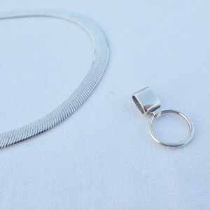 May include: A silver chain necklace with a silver pendant featuring a round ring. The chain is a herringbone style.