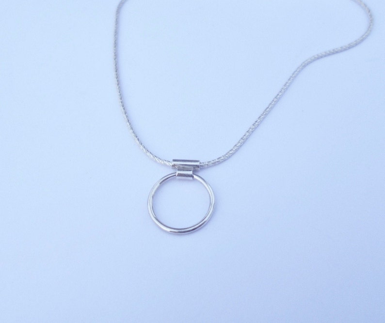 Pode incluir: Um colar de tom prateado com uma corrente fina e texturizada e um pingente circular simples, preso por um pequeno conector met&aacute;lico cil&iacute;ndrico. O design &eacute; uniforme, apresentando um acabamento met&aacute;lico em toda a pe&ccedil;a.