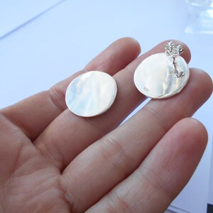 May include: A pair of silver round earrings with a smooth, polished finish. The earrings are shown on a hand, with the back of the earrings visible.