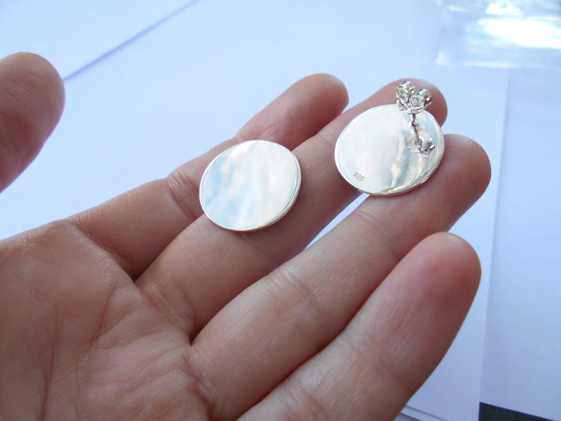 May include: A pair of silver round earrings with a smooth, polished finish. The earrings are shown on a hand, with the back of the earrings visible.