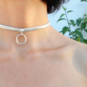May include: Silver choker necklace with a circular pendant. The necklace has a flat, woven design and sits around the neck. The pendant is a simple, open circle. The jewelry is displayed against a neutral background.