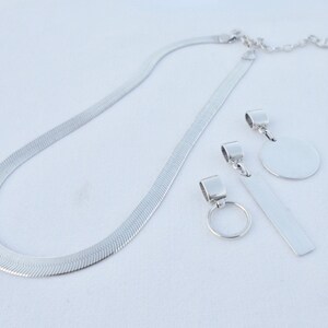 May include: A silver chain necklace with three silver pendant charms. The charms are a circle, a rectangle, and a small ring.