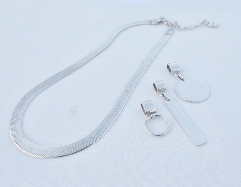 May include: A silver chain necklace with three silver pendant charms. The charms are a circle, a rectangle, and a small ring.