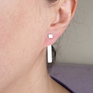 May include: A pair of silver earrings. One is a small square stud, and the other is a long, rectangular bar that hangs below the earlobe. The earrings are worn on a person's ear, with the focus on the jewelry.