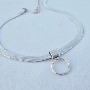 May include: A silver chain necklace with a herringbone pattern and a round pendant. The pendant is attached to the chain with a small, rectangular piece of metal.