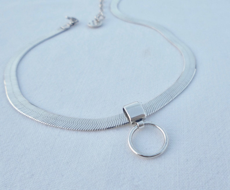 May include: A silver chain necklace with a herringbone pattern and a round pendant. The pendant is attached to the chain with a small, rectangular piece of metal.