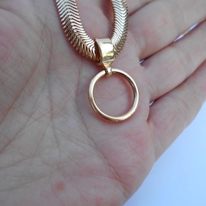 May include: A gold-colored necklace with a herringbone chain and a circular pendant. The necklace is displayed on a hand, showcasing the jewelry's design and color. The pendant is a simple, elegant circle.