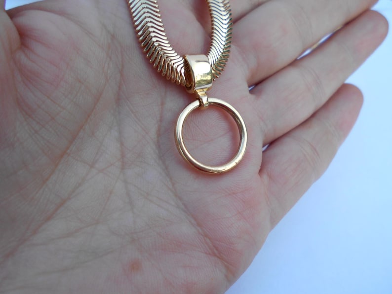 May include: A gold-colored necklace with a herringbone chain and a circular pendant. The necklace is displayed on a hand, showcasing the jewelry's design and color. The pendant is a simple, elegant circle.