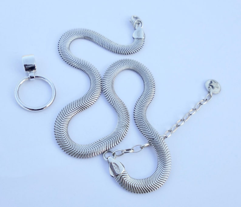 May include: A silver-colored necklace with a herringbone chain design, and a separate pendant with a circular loop. The necklace has a lobster clasp and an adjustable chain. The jewelry is displayed on a white background.