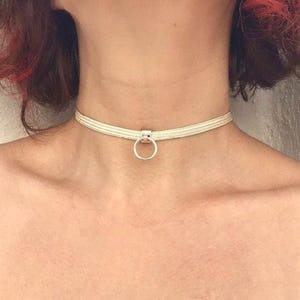 Può includere: Una collana girocollo argentata con un pendente circolare. La collana è realizzata con più fili di un materiale intrecciato di colore chiaro. Il pendente è un anello semplice e lucido. La collana è indossata intorno al collo.