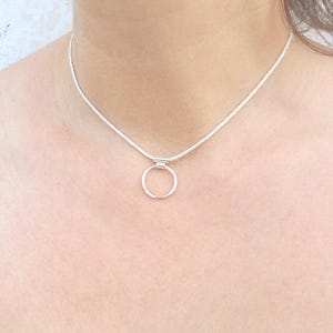 Pode incluir: Um colar de tom prateado com uma corrente de cobra fina e lisa e um &uacute;nico pendente de anel circular, fixado a um pequeno conector met&aacute;lico retangular no centro. O colar repousa sobre o pesco&ccedil;o, exibindo um acabamento met&aacute;lico limpo.