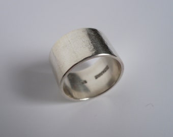 May include: A silver band ring with a smooth, flat surface. The ring is wide and has a rounded edge.