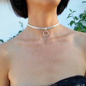 May include: A silver choker necklace with a small, round pendant. The necklace is a simple, elegant design, perfect for everyday wear or special occasions. The choker is made of a flat, woven material.