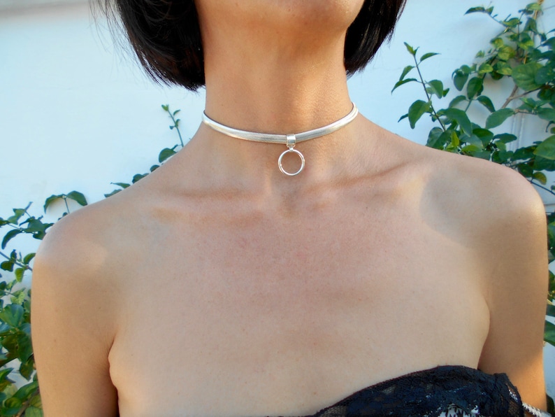 May include: A silver choker necklace with a small, round pendant. The necklace is a simple, elegant design, perfect for everyday wear or special occasions. The choker is made of a flat, woven material.