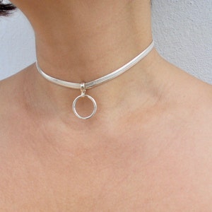 May include: A silver-toned snake chain choker necklace featuring a flat, flexible band design. The necklace includes a central circular ring pendant attached to the chain by a small, polished silver-toned bail, resting against the neck.