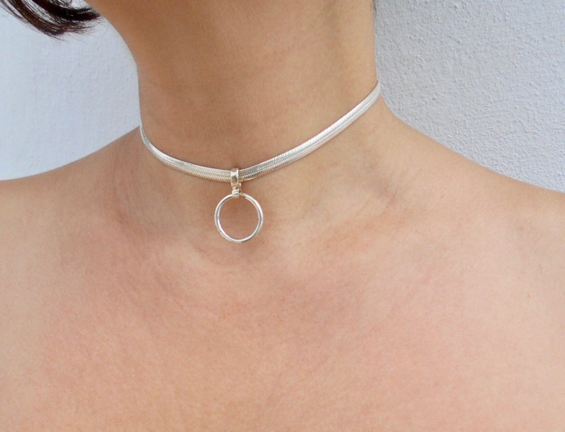 May include: A silver-toned snake chain choker necklace featuring a flat, flexible band design. The necklace includes a central circular ring pendant attached to the chain by a small, polished silver-toned bail, resting against the neck.
