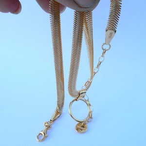 May include: A gold-colored herringbone chain necklace with a lobster clasp and a small charm. The necklace is displayed against a light blue background. The chain is held up by a hand.