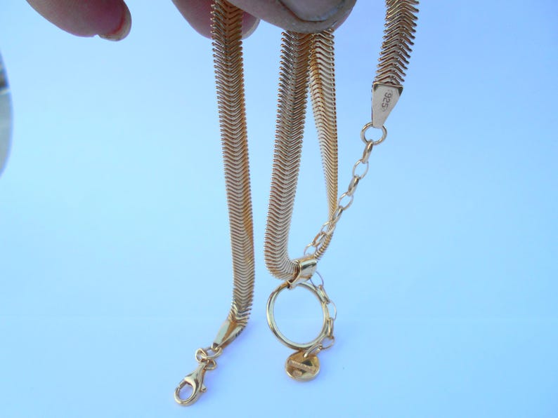 May include: A gold-colored herringbone chain necklace with a lobster clasp and a small charm. The necklace is displayed against a light blue background. The chain is held up by a hand.