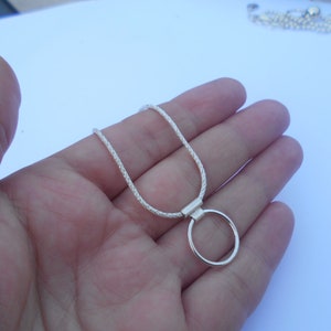 Pode incluir: Um colar de tom prateado com uma corrente texturizada estilo corda e um pingente circular liso preso por uma argola retangular. O colar tem um acabamento met&aacute;lico brilhante e um design simples que repousa na palma da m&atilde;o.