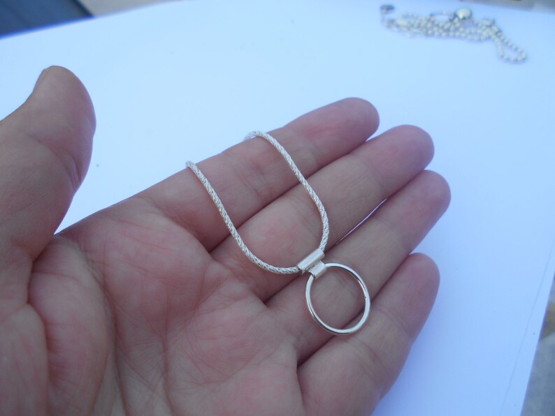 Pode incluir: Um colar de tom prateado com uma corrente texturizada estilo corda e um pingente circular liso preso por uma argola retangular. O colar tem um acabamento met&aacute;lico brilhante e um design simples que repousa na palma da m&atilde;o.