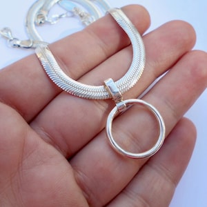 May include: A silver-toned herringbone chain necklace featuring a polished circular pendant attached to a rectangular bail. The necklace has a lobster claw clasp and a flat, woven texture that reflects light across its metallic surface.