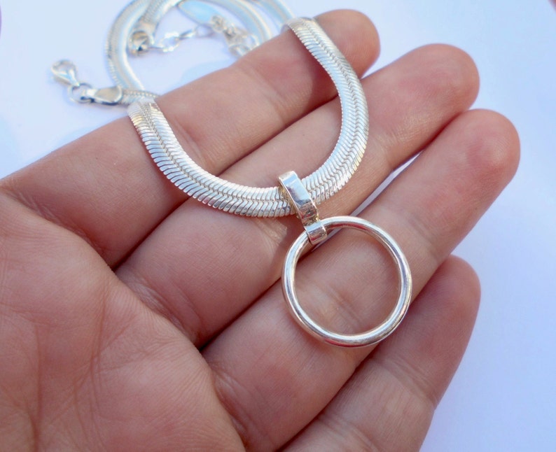 May include: A silver-toned herringbone chain necklace featuring a polished circular pendant attached to a rectangular bail. The necklace has a lobster claw clasp and a flat, woven texture that reflects light across its metallic surface.