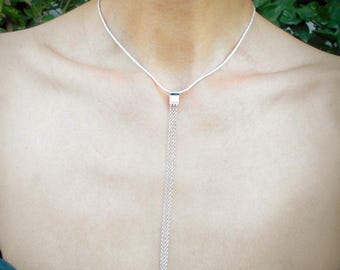 May include: A silver necklace with a delicate chain and a rectangular pendant. The pendant has a long, cascading fringe of fine chains. The necklace is worn against a person's skin, with a green top visible at the bottom of the frame.