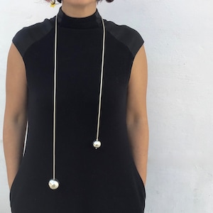 May include: A long silver chain necklace with two large silver ball pendants. The necklace is worn by a person wearing a black mock neck shirt.