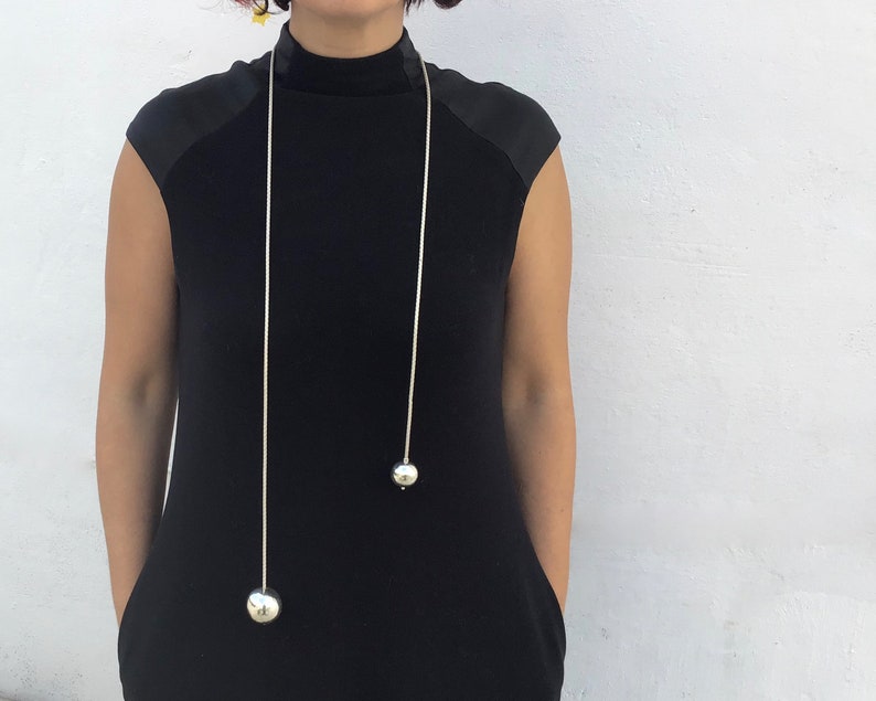 May include: A long silver chain necklace with two large silver ball pendants. The necklace is worn by a person wearing a black mock neck shirt.