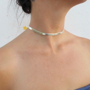 Pu&ograve; includere: Un girocollo in argento con una fascia piatta e larga. Il girocollo &egrave; indossato da una persona con i capelli scuri.
