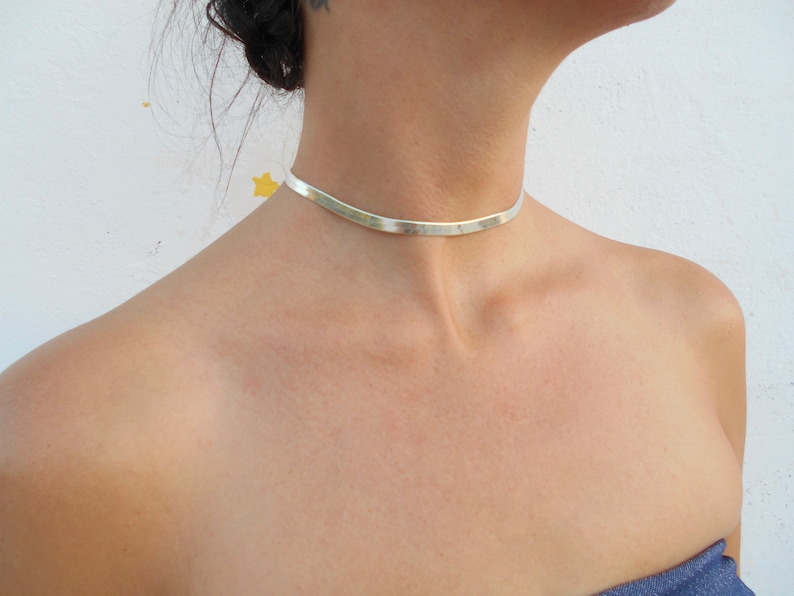 Pu&ograve; includere: Un girocollo in argento con una fascia piatta e larga. Il girocollo &egrave; indossato da una persona con i capelli scuri.