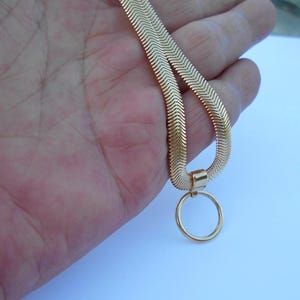 May include: A gold-colored herringbone chain necklace with a circular pendant. The necklace is held in a hand against a light blue background. The chain has a flat, chevron design.