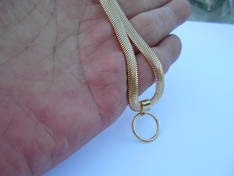 May include: A gold-colored herringbone chain necklace with a circular pendant. The necklace is held in a hand against a light blue background. The chain has a flat, chevron design.