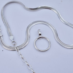 May include: A silver-colored herringbone chain necklace featuring a lobster claw clasp and an adjustable extender chain with oval links. A circular pendant is positioned separately next to the chain, featuring a smooth, polished silver-colored finish.