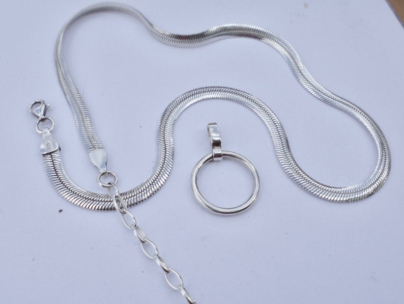 May include: A silver-colored herringbone chain necklace featuring a lobster claw clasp and an adjustable extender chain with oval links. A circular pendant is positioned separately next to the chain, featuring a smooth, polished silver-colored finish.