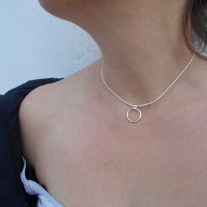 Pode incluir: Um colar tipo gargantilha de tom prateado com uma corrente fina e texturizada e um &uacute;nico pingente de anel circular liso centrado na frente. O colar repousa rente ao pesco&ccedil;o, exibindo um design met&aacute;lico simples com acabamento polido.
